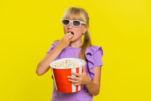 Excited young cute school girl in 3D glasses eating popcorn, watching interesting tv serial, sport game, film, online social media movie content. Teen female child kid on studio yellow background