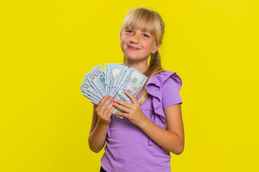 Rich pleased young cute school girl waving money dollar cash banknotes bills like a fan, success, lottery winner, big income, wealth. Teen blonde female child kid on studio yellow background
