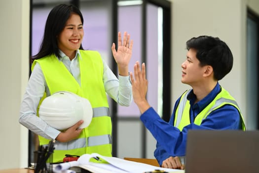 Smiling asian female engineer giving high five with colleague, celebration for teamwork achievement.