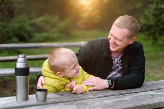 Happy family: father and child boy son playing and laughing in autumn park, sitting on wooden bench. Father and little kid having fun outdoors, playing together. Father and son sitting on a bench and talking. dad son park bench table autumn thermos.