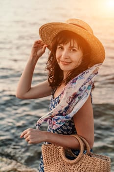 A woman in a dress, hat and with a straw bag is standing on the beach enjoying the sea. Happy summer holidays.