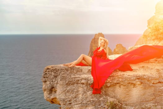 woman red silk dress sits by the ocean with mountains in the background, her dress swaying in the wind