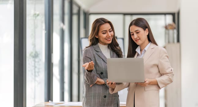 Two happy professional coworkers discussing plan at work. explaining Asian colleague financial project on laptop in office.