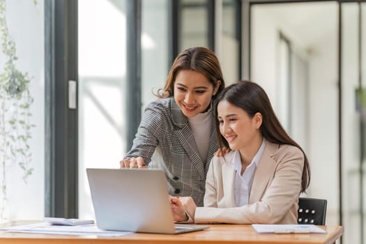 Two happy professional coworkers discussing plan at work. explaining Asian colleague financial project on laptop in office.