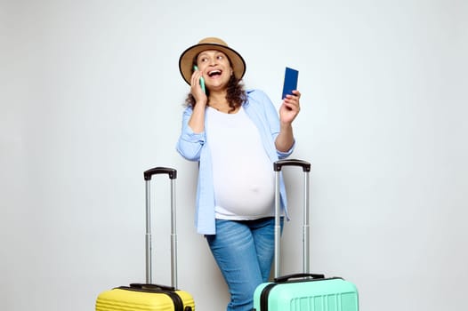 Happy smiling pregnant woman traveler talking on mobile phone, booking hotel, posing with boarding pass and suitcases, isolated on white background. The concept of safe travel and tourism in pregnancy