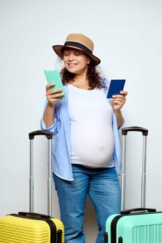 Happy pregnant woman checking her flight on mobile app using her smartphone, going on summer vacations, standing with a boarding pass and suitcases, smiling cutely, isolated on white studio background