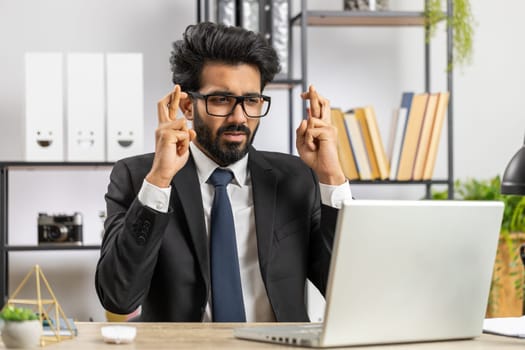 Bearded indian businessman crossed fingers looking at laptop screen asking for good luck news, wishing good exam results, dreaming about win victory in lottery jackpot, business money profit at office