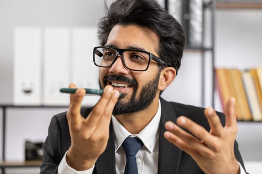 Bearded indian businessman working, having mobile phone loudspeaker talk at office desk with laptop. Manager freelance indian man holding smartphone using messenger chat apps. Employment, occupation