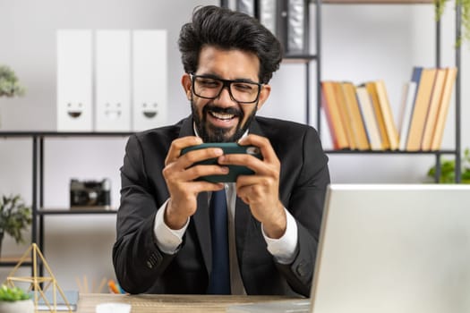 Indian businessman playing on smartphone exciting mobile application celebrate winning racing or shooter game at office desk workplace. Professional manager freelancer man enjoy good work of new app