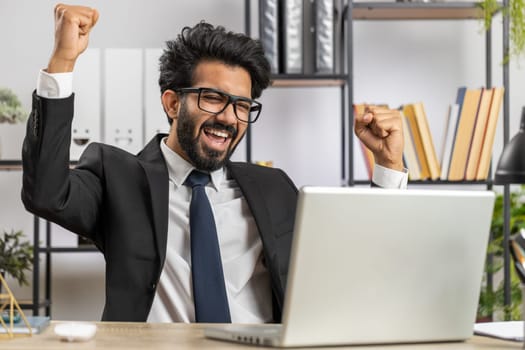 Indian businessman working on laptop at home office desk typing browsing say Wow yes found out great big lottery jackpot win, good job news, celebrating. Manager freelancer arab man