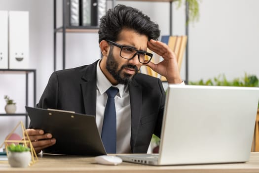 Irritated tired indian business man while working on laptop, unexpected online website problem, computer virus data loss by hacking. Freelancer feeling mad about broken notebook at office workplace