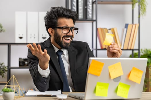 Tired exhausted indian businessman working on laptop at office with many sticker task inscriptions. Freelancer man busy occupation feels sad boredom overworked multitasking. Panic attack, deadline