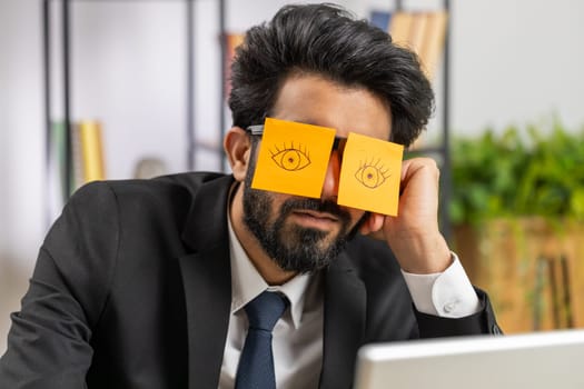 Inefficient tired young businessman working sleeping on laptop computer with eyes stickers on face at office workplace desk. Indian lazy manager freelancer man. Business people cheating to sleep