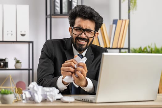 Angry furious indian man working at office throwing crumpled paper, having nervous breakdown at work, migaine, headache, stress management, mental distress problems, losing temper, reaction on failure
