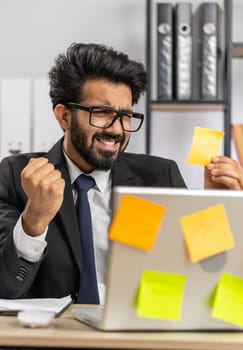 Tired exhausted indian businessman working on laptop at office with many sticker task inscriptions. Freelancer man busy occupation feels sad boredom overworked multitasking. Panic attack, deadline