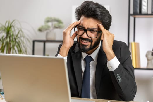 Tired ill indian businessman suffering from headache problem tension and migraine, stress at home office desk workplace. Bearded freelancer broker worker man works on laptop notebook. Overworking