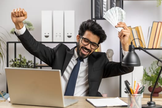 Indian businessman holding fan of cash money dollar banknotes celebrate, dance, success career, lottery jackpot game winner, big income, wealth at home office workplace desk. Remote distant working