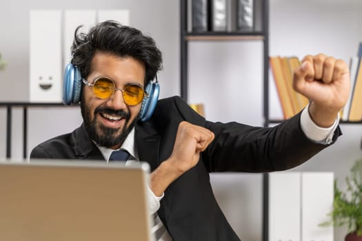 Happy relaxed overjoyed indian businessman working on laptop computer at home office wearing headphones listening favorite energetic disco music and dancing. Freelancer man relaxing, taking a break