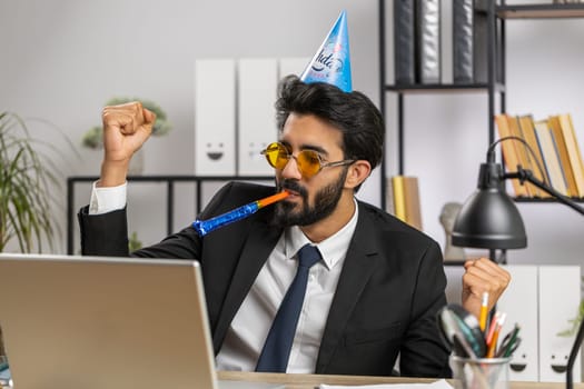 Excited lucky business man in sunglasses, festive cap hat cone blow pipe at workplace dancing victory dance, weekend. Rejoicing developing project. Celebrating birthday. Lottery jackpot. Office people