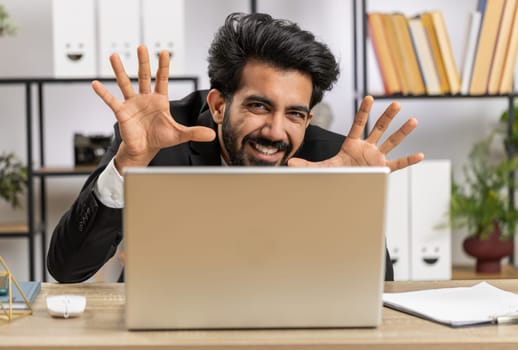 Indian businessman hiding behind laptop computer, making funny face, bullying, abuse, fooling around, remote working disrespecting someone at office desk workplace. Professional freelancer man