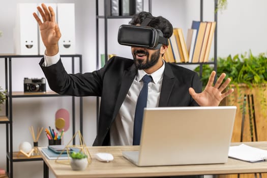 Young indian businessman programmer software developer using headset helmet app to play simulation game. Watching virtual reality 3D 360 video. Young hindu man in VR goggles working at home office