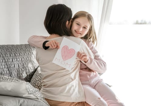 Cute little girl hugging mother and showing card with painted heart. Mother's day concept