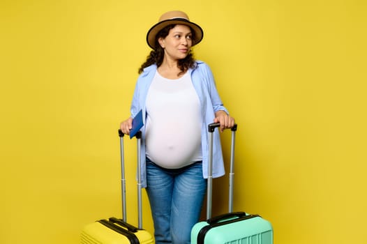 Multi-ethnic charming pregnant woman with stylish color suitcases, travelling abroad, going for weekend getaway, looking aside a copy space, isolated on yellow studio background. Travel and pregnancy