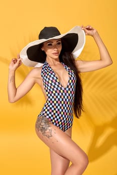 Photo of young beautiful brunette girl posing in the studio, standing in a black-and-white hat and checkered swimsuit. Yellow background