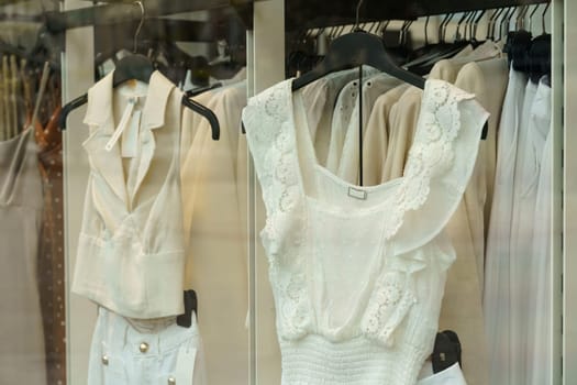 Light-colored women's clothing hangs on display racks behind glass in a shop window.