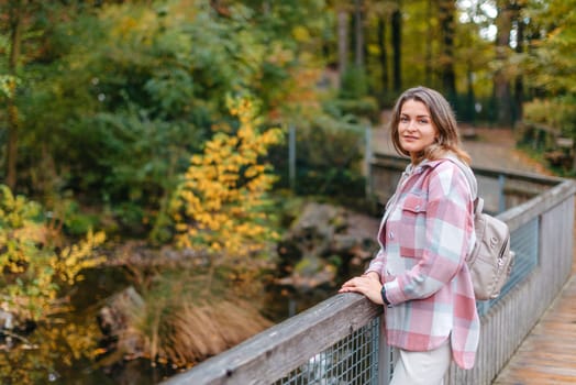 Portrait of cute young woman in casual wear in autumn, standing on bridge against background of an autumn Park and river. Pretty female walking in Park in golden fall. Copy space. smiling girl in the park standing on wooden bridge and looking at the camera in autumn season