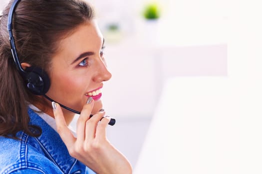 Happy charming young woman sitting and working with laptop using headset in office.