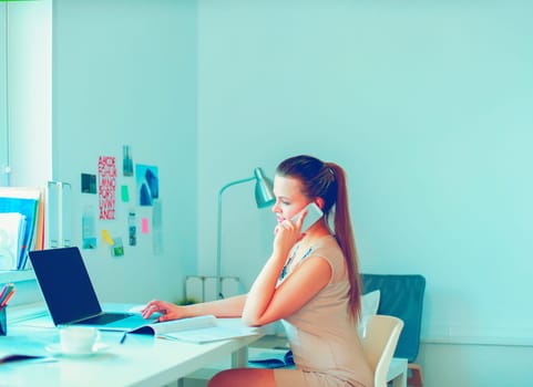 Attractive woman sitting at desk in office, working with laptop computer, holding document.