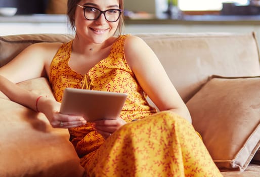 Beautiful young brunette woman at home sitting on sofa or settee using her tablet computer and smiling.