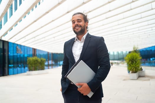 Successful businessman in suit with beard standing in front of office building confidently looking at camera and smile holding document. Hispanic male business person. Side view portrait