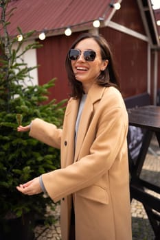 Woman looking for perfect Christmas tree to buy on the tree shop, outdoor in European city. Girl using hand for check the quality. Dressed stylish trench coat and sunglasses