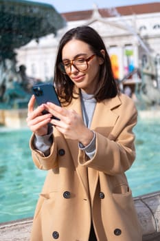 Woman wearing glasses, sitting at the fountain, holding smartphone and smiling sweetly. Good vibes and feelings. Online communication using mobile digital device