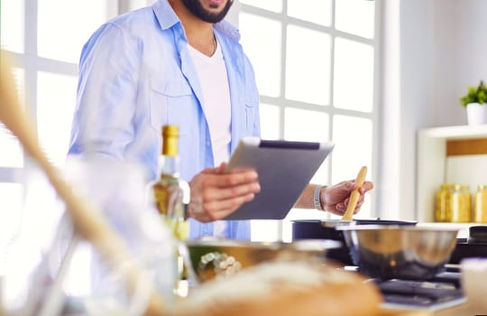 Man following recipe on digital tablet and cooking tasty and healthy food in kitchen at home.