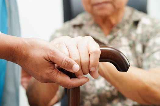 Doctor help Asian elderly disability woman patient holding walking stick in wrinkled hand at hospital.