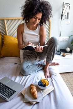 Young multiracial latina woman writing daily affirmations on journal while eating breakfast in bed next to laptop. Ready to work. Vertical image. Lifestyle concept.