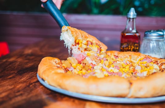Person hand taking a slice of sweetcorn and ham pizza with spatula. Delicious ham pizza with cheese and sweet corn
