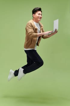 Full length portrait of a happy young man jumping with laptop computer isolated over green background