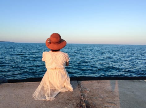a girl in a white dress and a hat sits on the pier and looks at the sunset sea, rear view, copy space