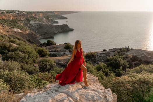 Woman sunset sea red dress, side view a happy beautiful sensual woman in a red long dress posing on a rock high above the sea on sunset