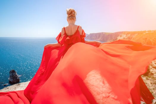 woman sea red dress. Blonde with long hair on a sunny seashore in a red flowing dress, back view, silk fabric waving in the wind. Against the backdrop of the blue sky and mountains on the seashore