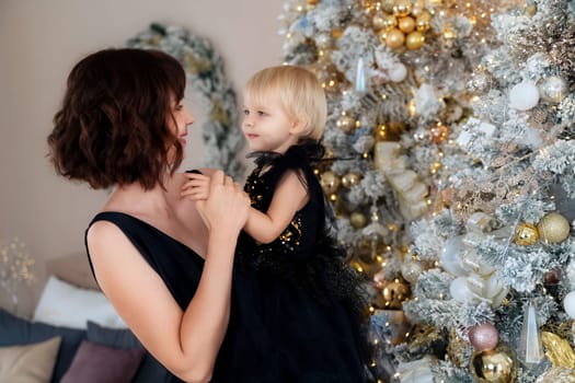 Mother with her daughter 2 years old near the Christmas tree. Both are dressed in black dresses, the mother holds the girl in her arms and both look at each other. The family celebrates Christmas