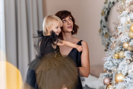 Mother daughter 2 years old Christmas tree. Both are dressed in black dresses, the mother holds the girl in her arms and both look at each other. The family celebrates Christmas