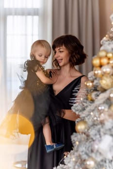 Mother daughter 2 years old Christmas tree. Both are dressed in black dresses, the mother holds the girl in her arms and both look at each other. The family celebrates Christmas