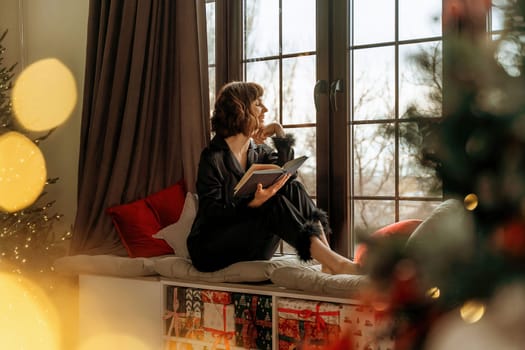 Woman christmas window. A beautiful brunette woman in black pajamas sits on the windowsill near the Christmas tree and looks out the window with a book in her hand, the room is decorated for Christmas. Holiday at home