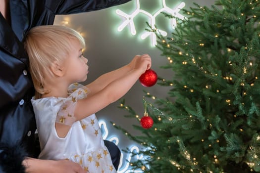 A mother with a 2-year-old daughter decorates the Christmas tree. Mom in a black suit, a girl in a white dress, her daughter hangs a red ball on the Christmas tree. Merry Christmas and New Year concept.