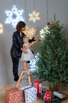 A mother with a 2-year-old daughter decorates the Christmas tree. Mom in a black suit, a girl in a white dress, her daughter hangs a red ball on the Christmas tree. Merry Christmas and New Year concept.